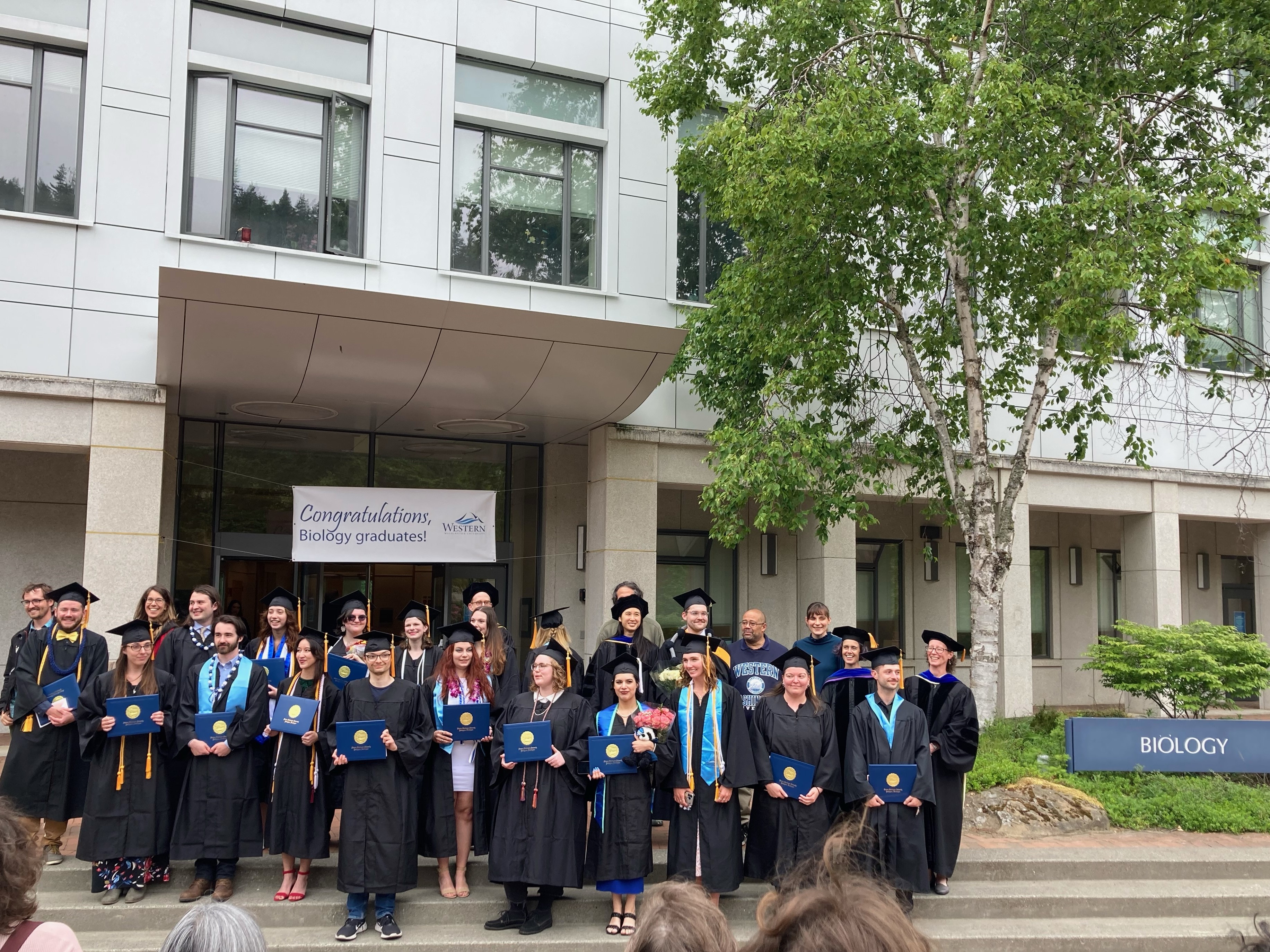 Graduates and faculty in caps and gowns stand in rows on the front steps of the biology building