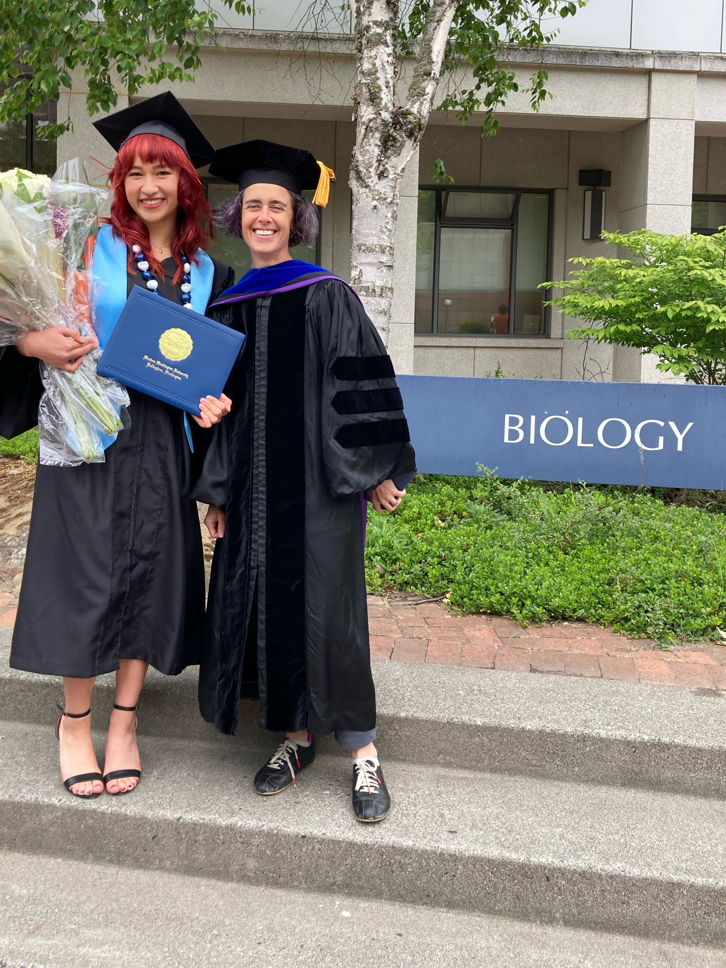 Graduate and faculty member in cap and gown stand in front of the Biology building sign. Graduate is holding diploma and flowers.
