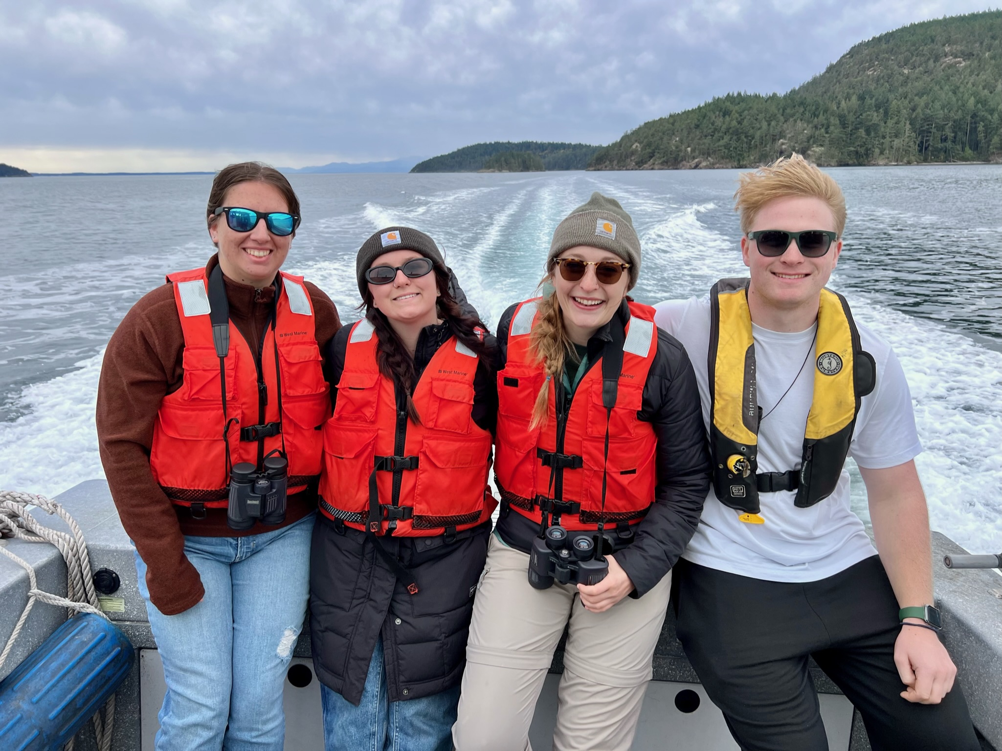 Four student researchers wearing bright life vests ride in a boat. Islands are visible in the background