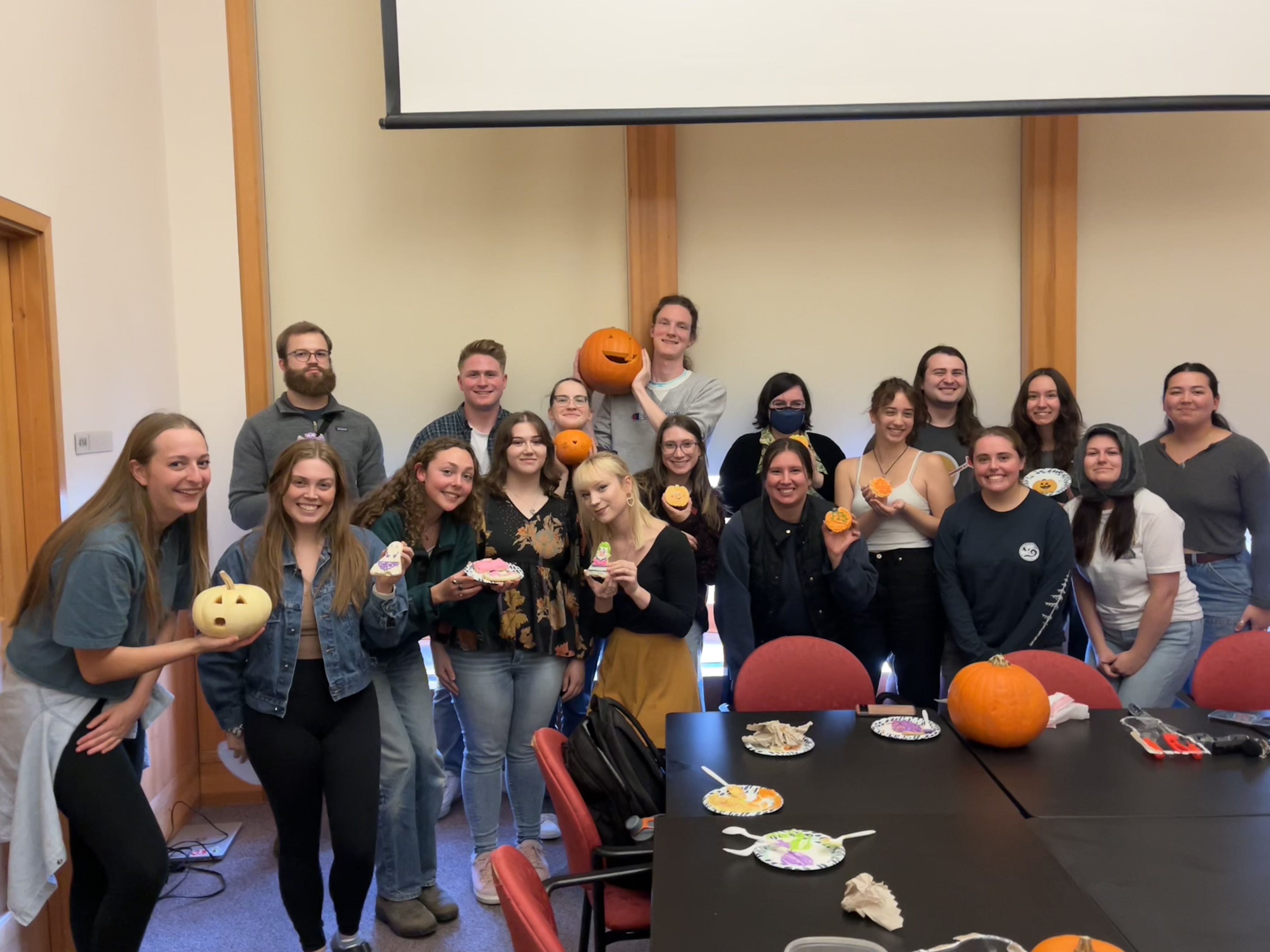 Large group of students in a meeting room hold pumpkins that have been carved and cookies that have been decorated at a celebration.
