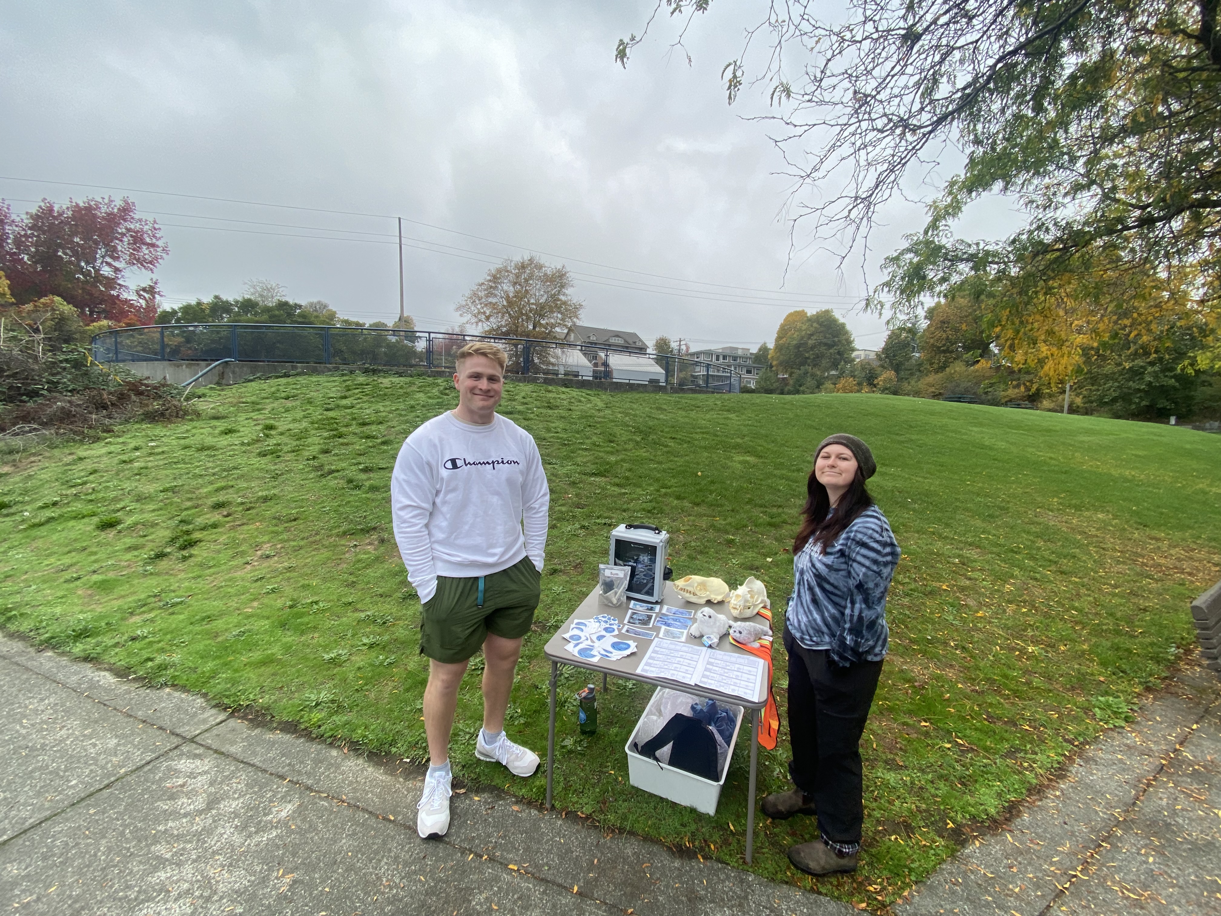 Two students stand on the edge of the sidewalk by a grassy field. The table between them has informational pamphlets.