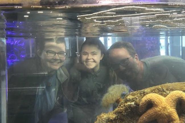 Three people stand on opposite of aquarium tank looking through the water and glass smiling at the camera.